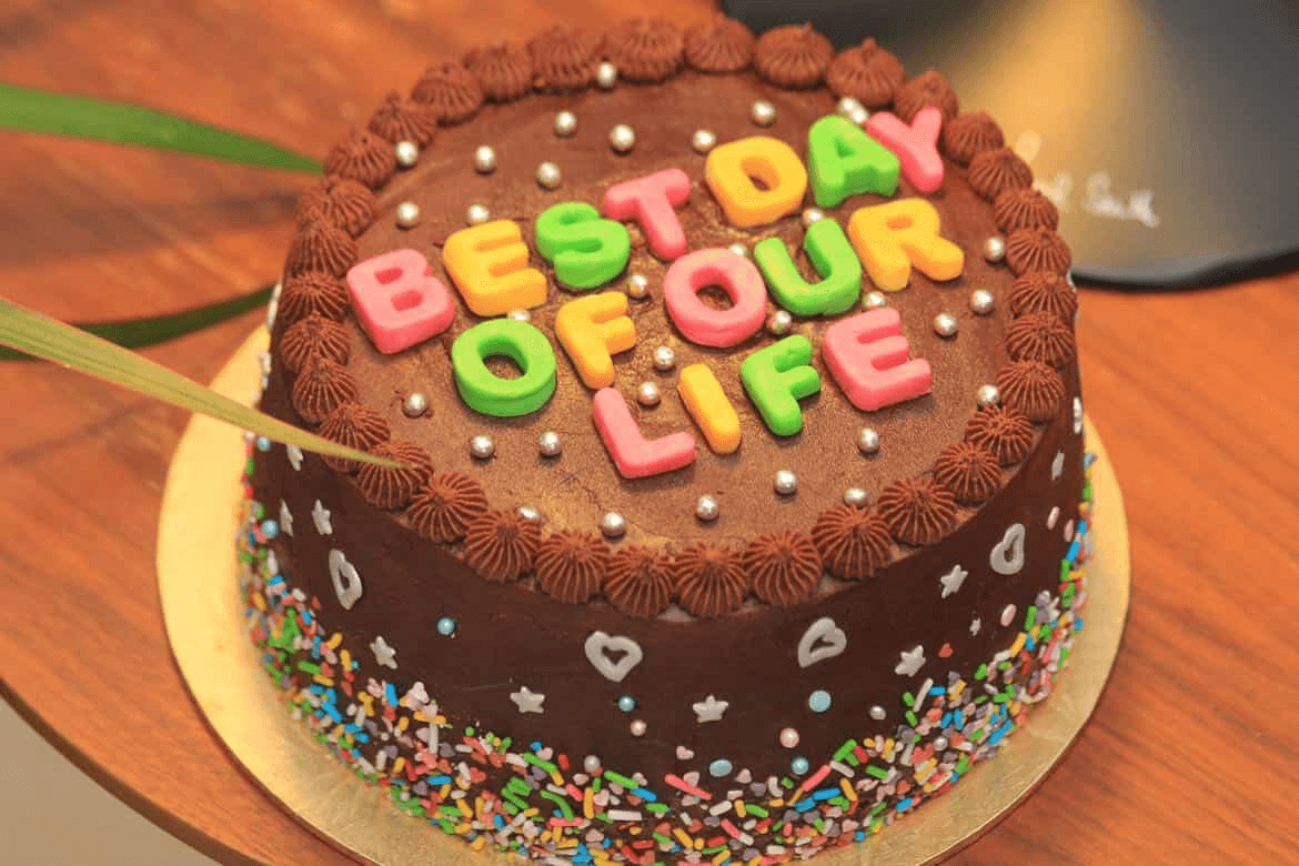 Round chocolate buttercream cake on a gold board, ringed with piped chocolate stars and scattered with silver dragees, with multicoloured fondant capitals across the top reading "BEST DAY OF OUR LIFE", and rainbow sprinkles, small silver hearts and pastel sugar pearls covering the sides