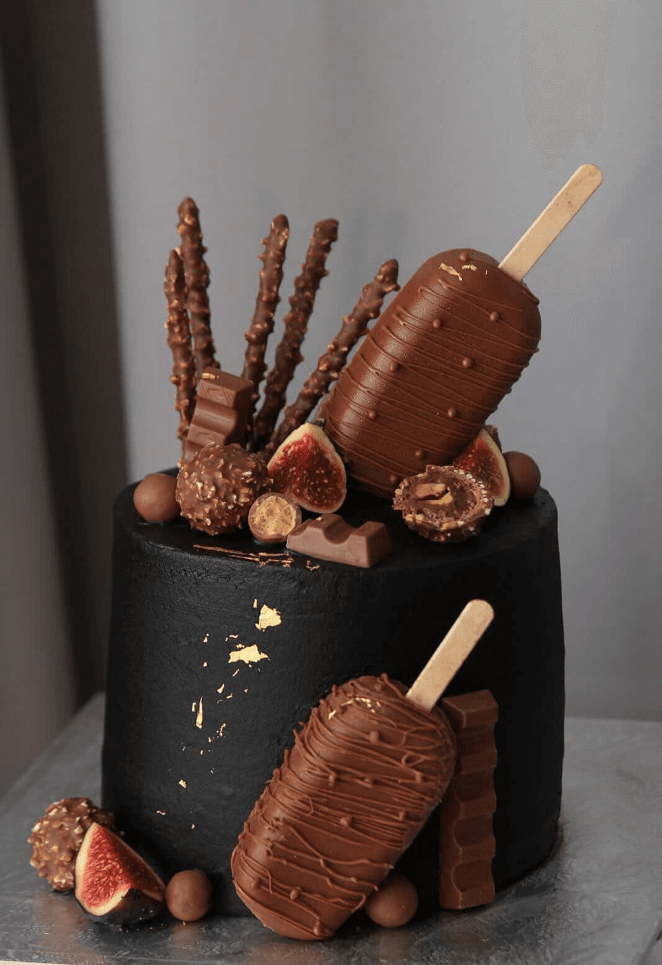 Tall cake in matte black icing flecked and streaked with gold leaf, topped with a chocolate-coated ice-cream bar standing on its stick, a fan of chocolate-dipped nut-studded wafer sticks, hazelnut truffles, malted chocolate balls, broken chunks of milk chocolate and halved fresh figs, with a second ice-cream bar, a wafer bar and more figs propped against the base