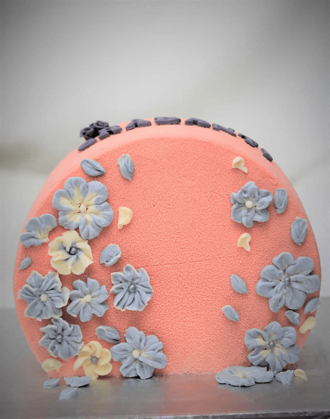 Round cake stood upright on its edge, finished in coral velvet, with hand-piped buttercream blossoms in dusty periwinkle blue and cream massed down both curved sides and thinning to loose petals across the bare centre, the flowers centred with silver pearls or piped cream, and a row of mauve piped shells and one piped mauve rose along the top curve