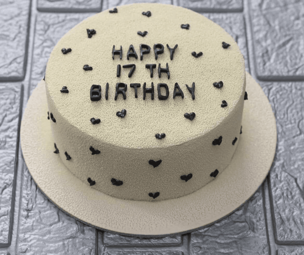 Pale sage-cream velvet cake on a wide matching covered board, scattered all over the top and sides with small black piped hearts, with "Happy 17th Birthday" lettered across the top in black, photographed against a grey brick-effect wall
