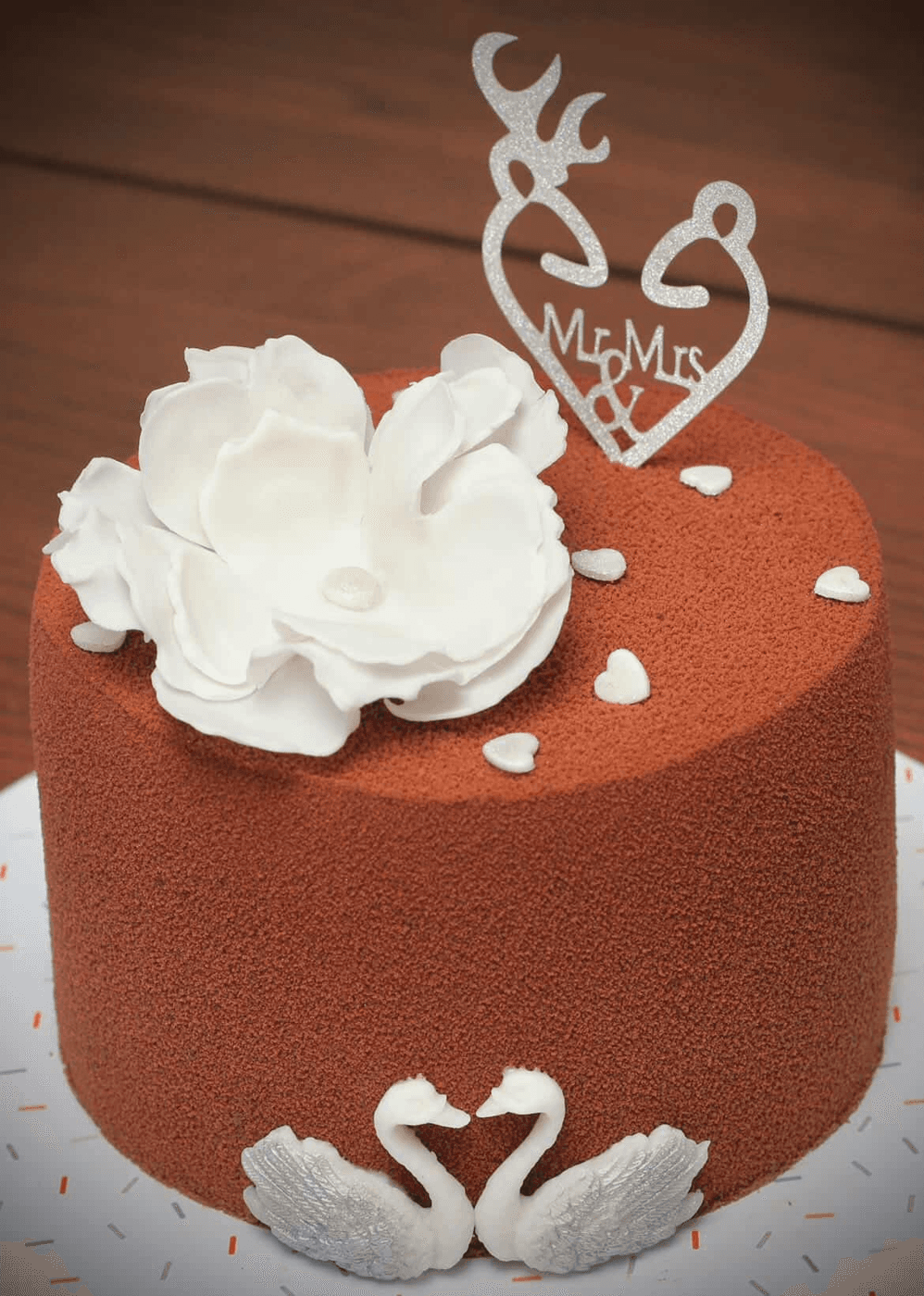 Single-tier cake in terracotta velvet, topped with one large white sugar magnolia and scattered small white sugar hearts, a pair of white sugar swans facing each other at the base, and a silver glitter topper cut as a stag and a doe whose heads and antlers form two interlocking hearts around the words Mr & Mrs, standing on a confetti-print board
