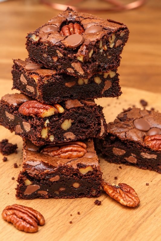 Stylised image of a stack of fudgy chocolate brownies studded with pecans and milk chocolate buttons on a wooden board.