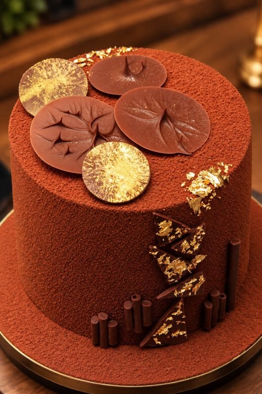 Stylised image of a cocoa velvet chocolate ganache cake topped with chocolate discs, gold leaf and a cascade of dark chocolate shards on a gold rimmed board.