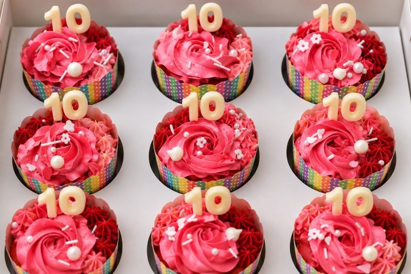 Stylised image of nine cupcakes topped with two-tone pink and red buttercream roses, sugar pearls and white chocolate toppers in a white box.